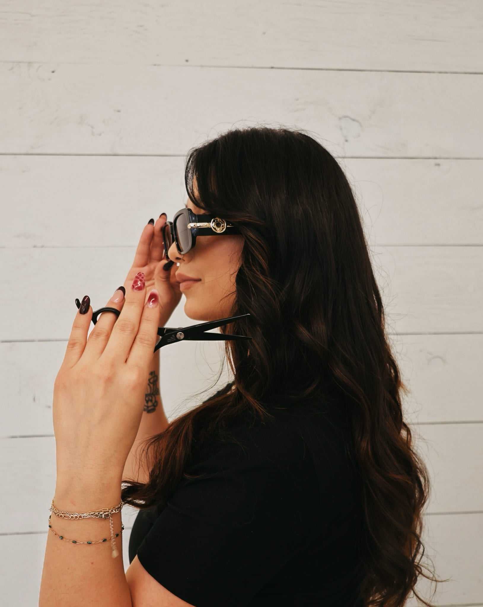 Woman with long hair holds sunglasses and scissors, facing a white wooden wall.