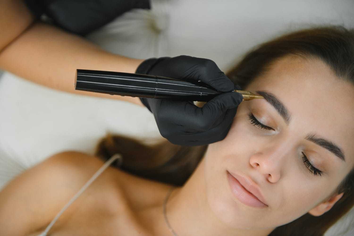 Woman receiving eyebrow microblading procedure by professional wearing gloves.