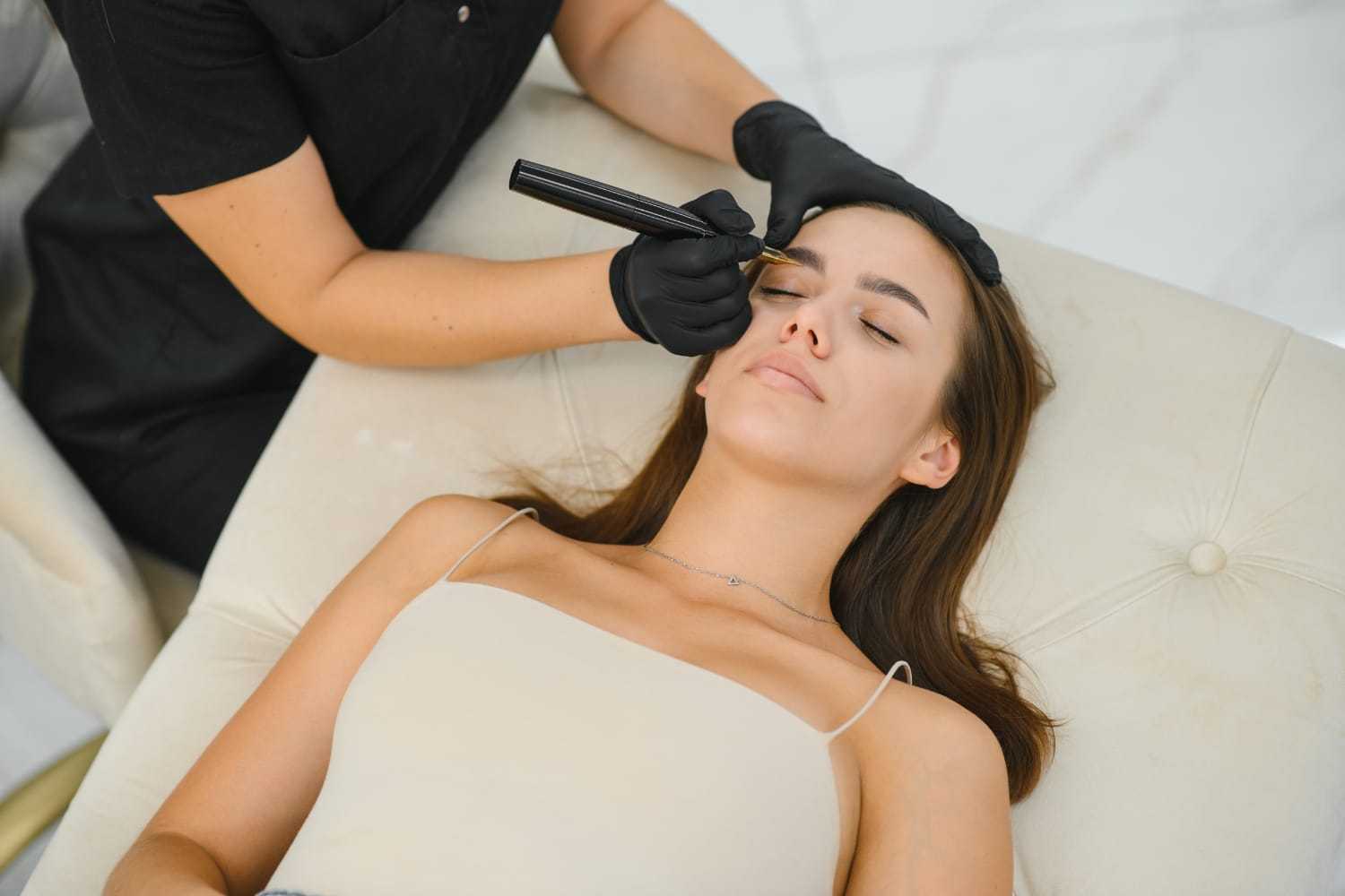 Woman receiving eyebrow treatment on a light-colored couch.