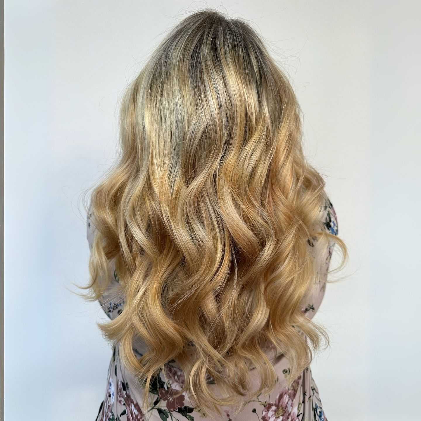 Woman with long, wavy blonde hair styled in soft curls wearing a floral patterned outfit.