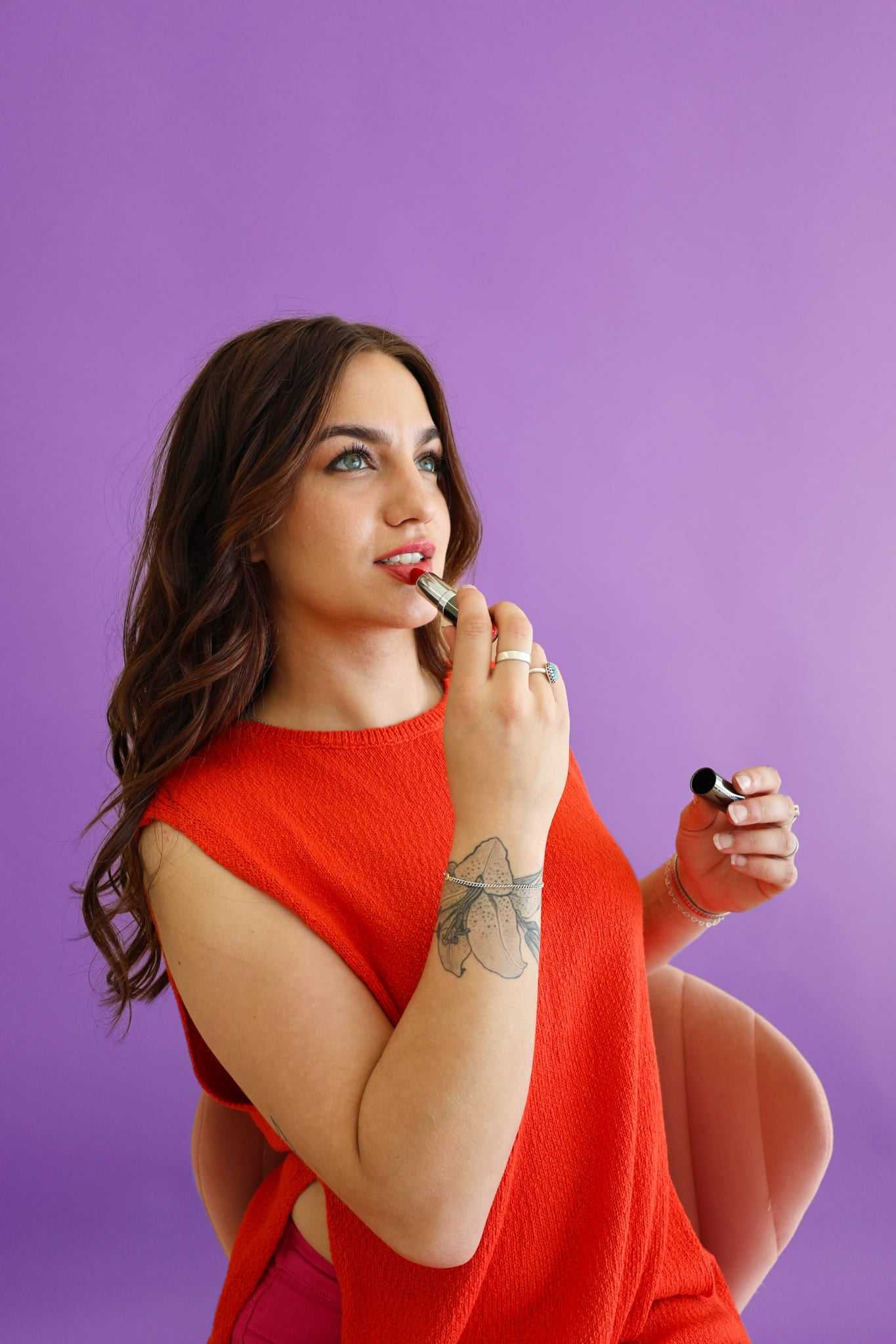 Young woman in red top applying lipstick against a purple background.