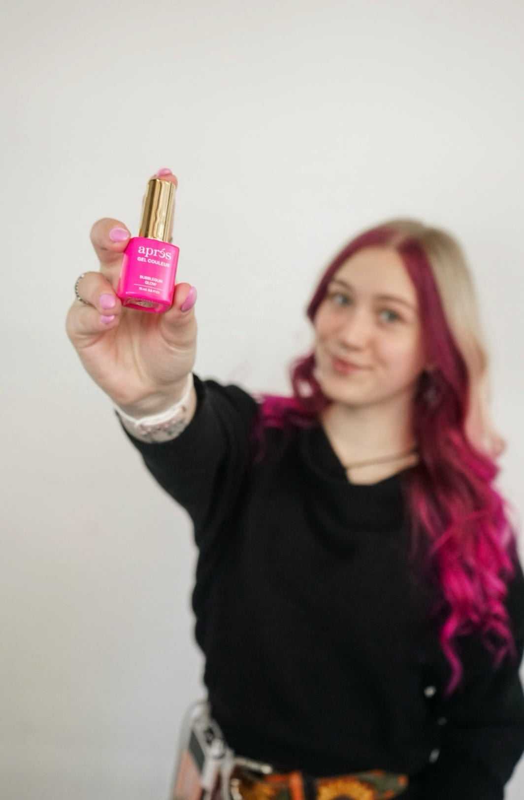 Person holding pink Aprisa bottle, smiling, with ombre hair against a plain background.