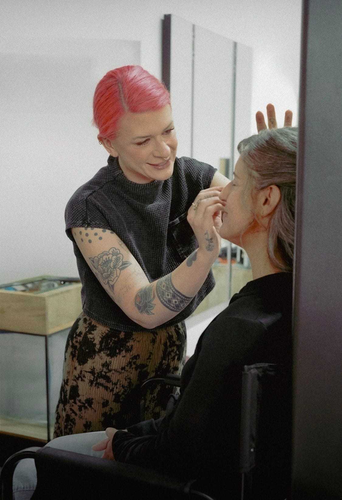 Makeup artist with tattoos applies makeup to a person's face in a studio setting.