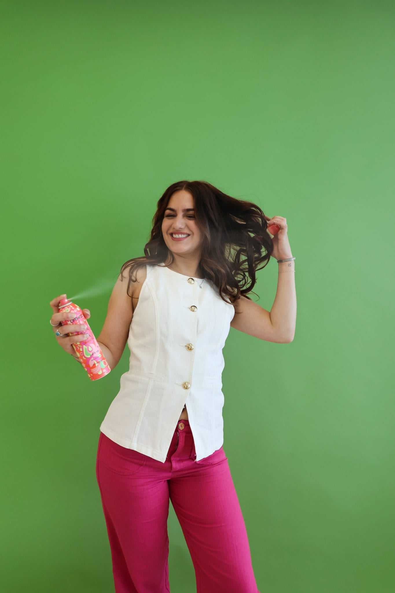 Woman in white top and pink pants sprays hair with can, standing against a green background.