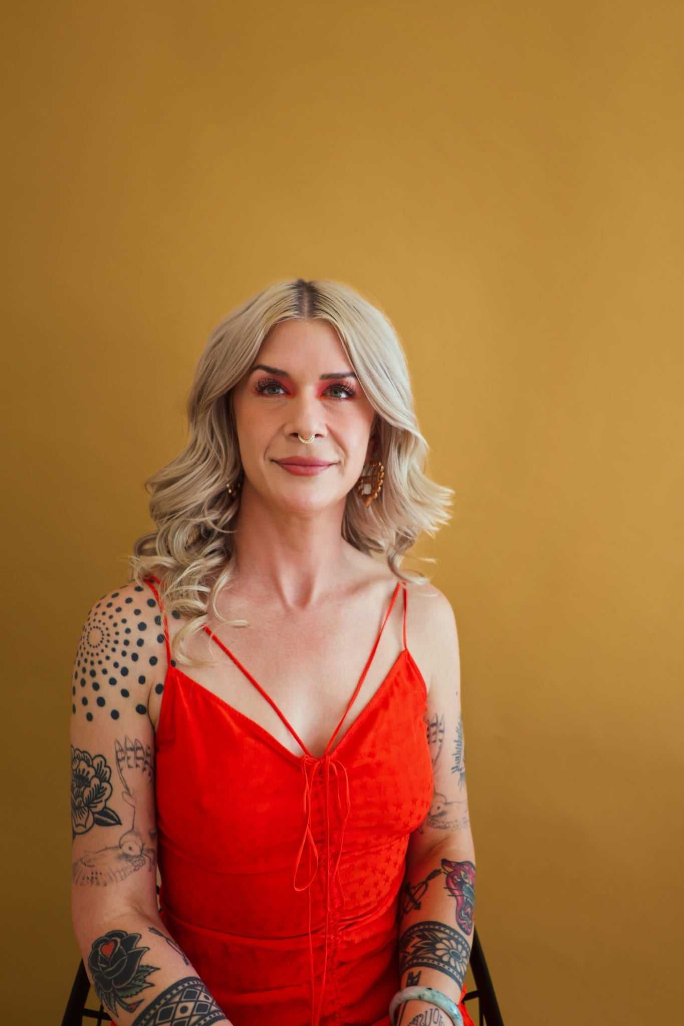Blonde woman in a red dress with tattoos, sitting against a mustard background.
