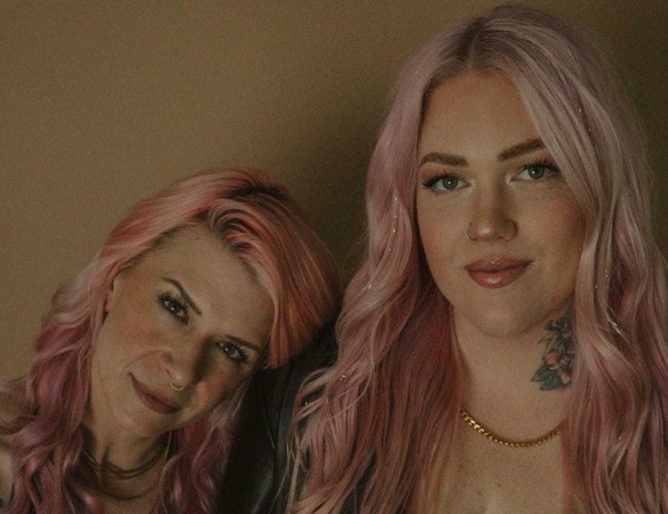 Two smiling people with pink hair posing together against a neutral background.