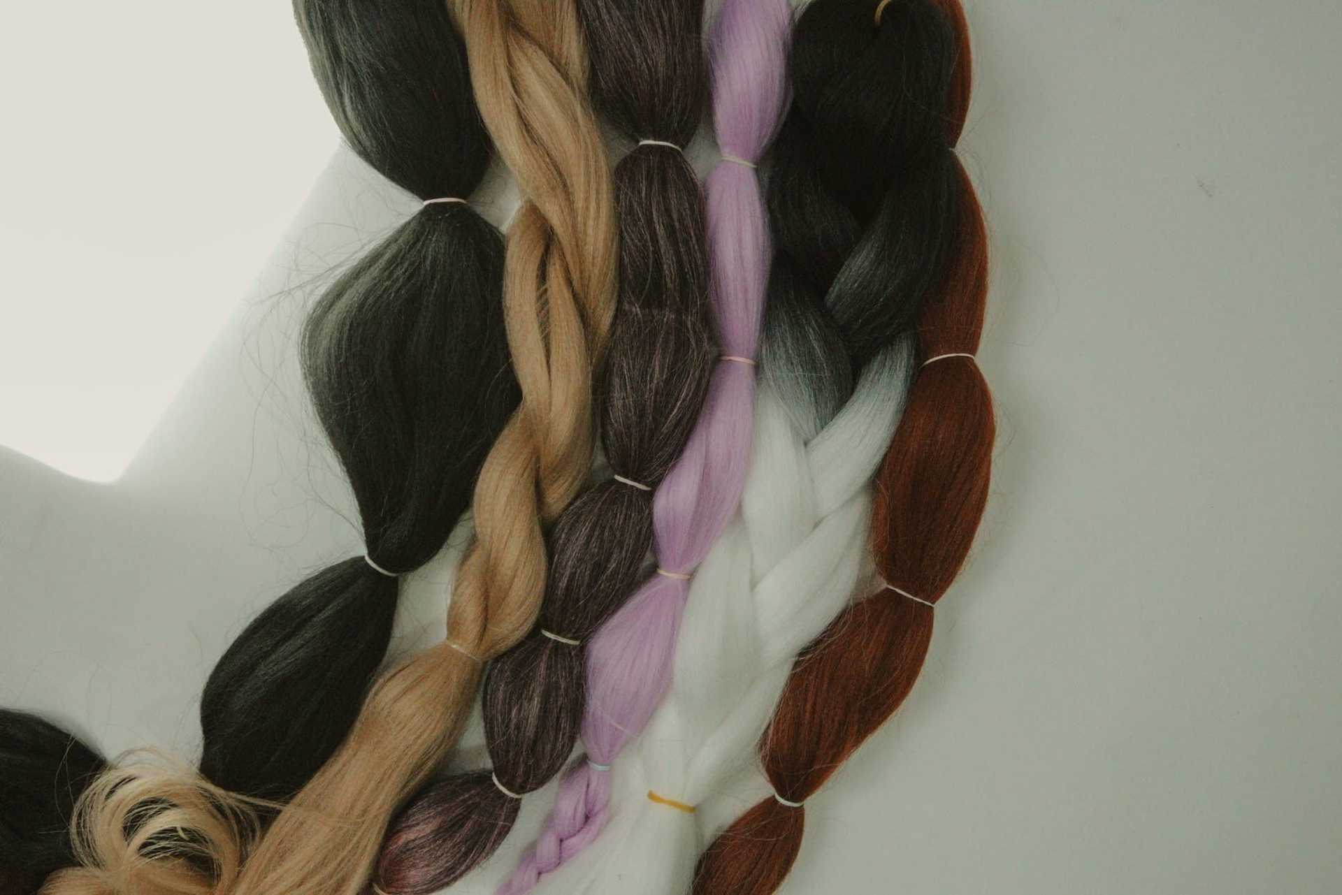 Braids of hair in various colors: black, brown, blonde, lavender, gray, and white.