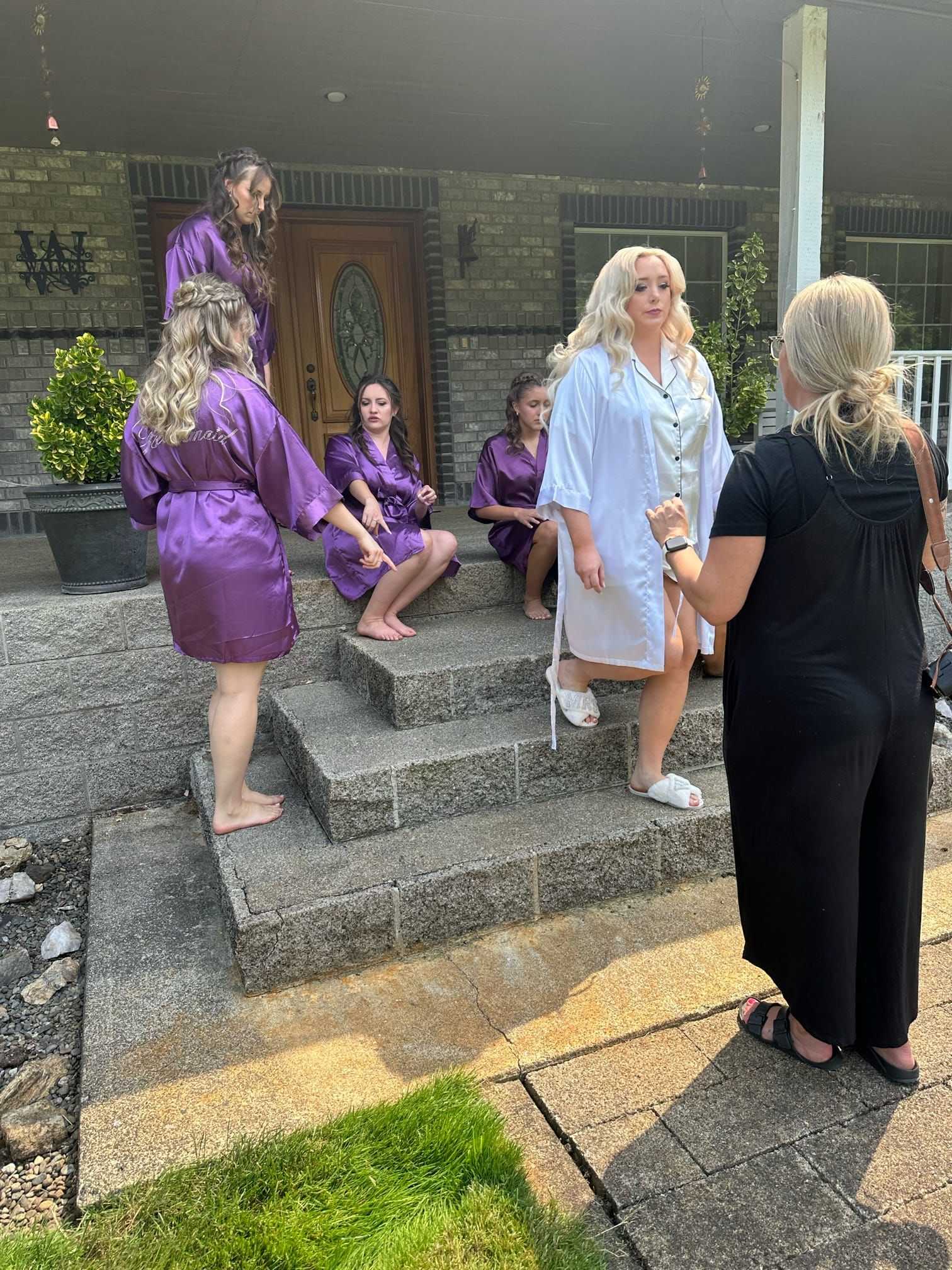 Bridesmaids in purple robes and bride in white robe talk on the steps of a brick house.