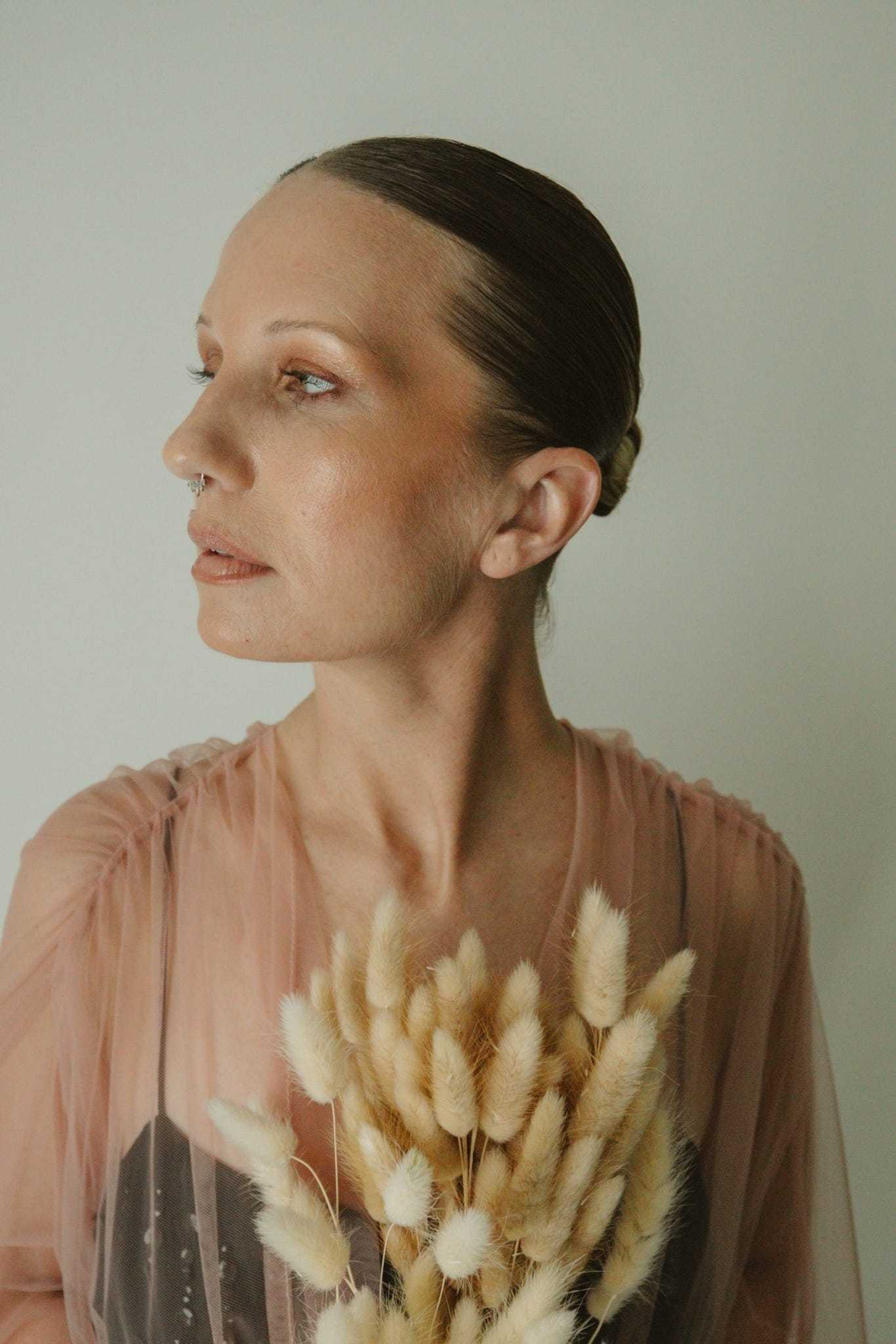 Woman holding dried flowers, gazing to the side, wearing sheer pink fabric.
