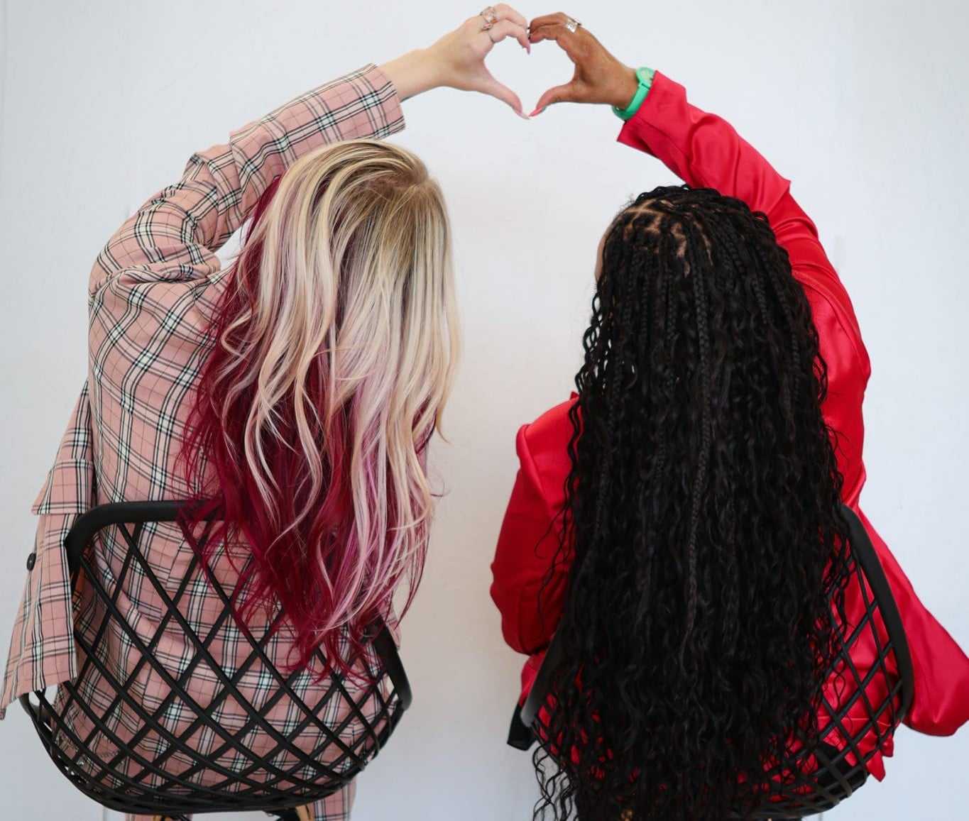 Two people with long hair form a heart shape with their hands, sitting back to back.