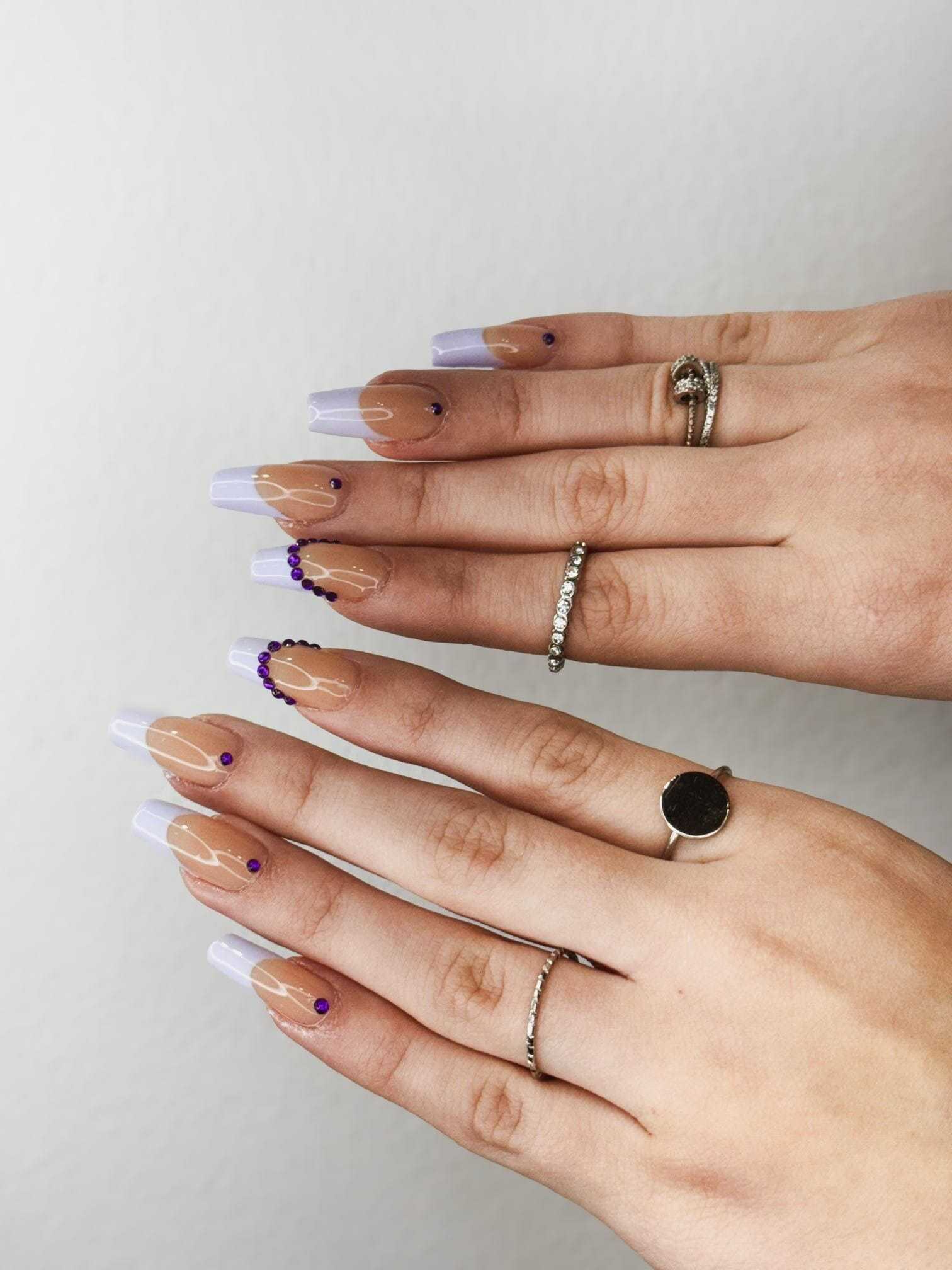 Hands with manicured nails, purple accents, and rings against a plain background.