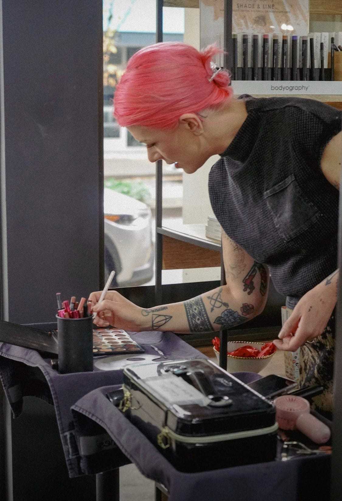 Makeup artist with pink hair applying cosmetics on a palette in a studio setting.