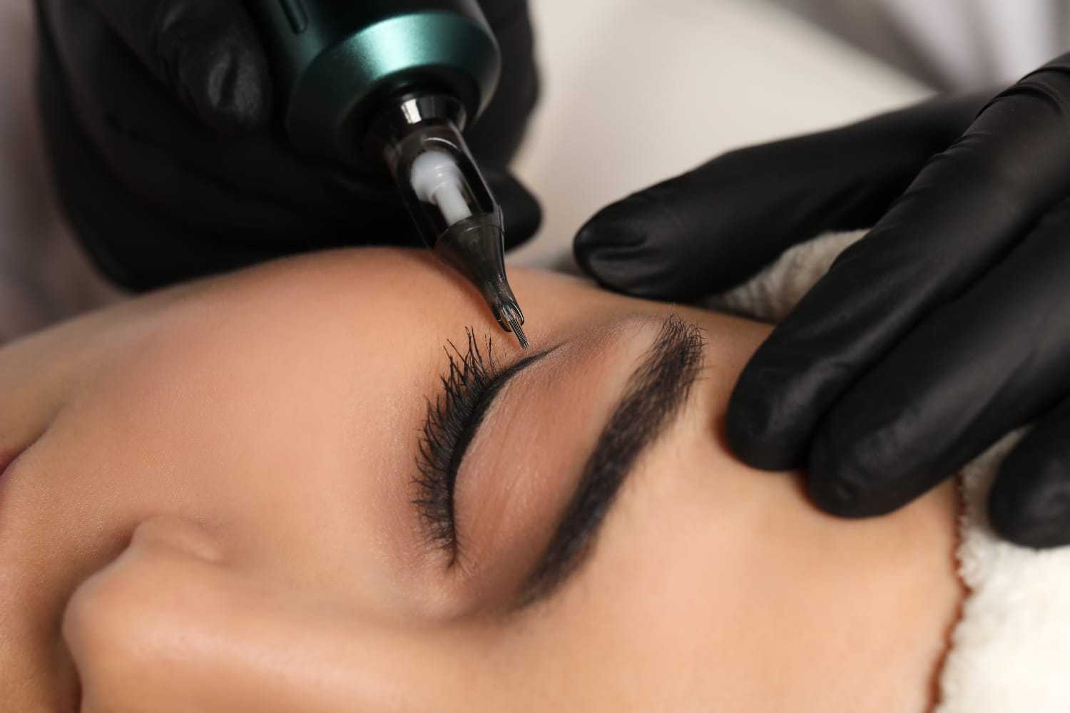 Eyebrow being tattooed by a professional with precision tools and gloves.