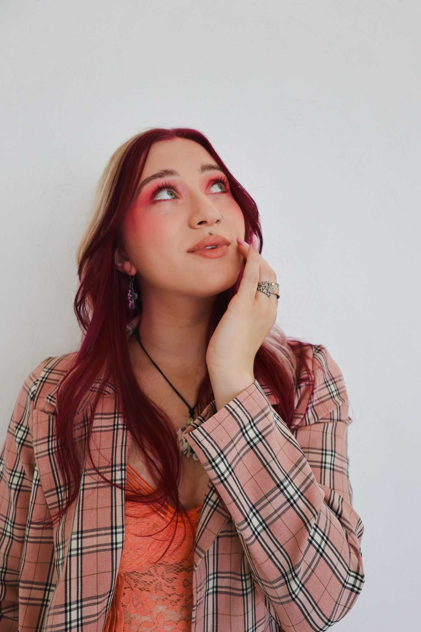 Person with red hair in pink plaid jacket, looking up thoughtfully against a white background.