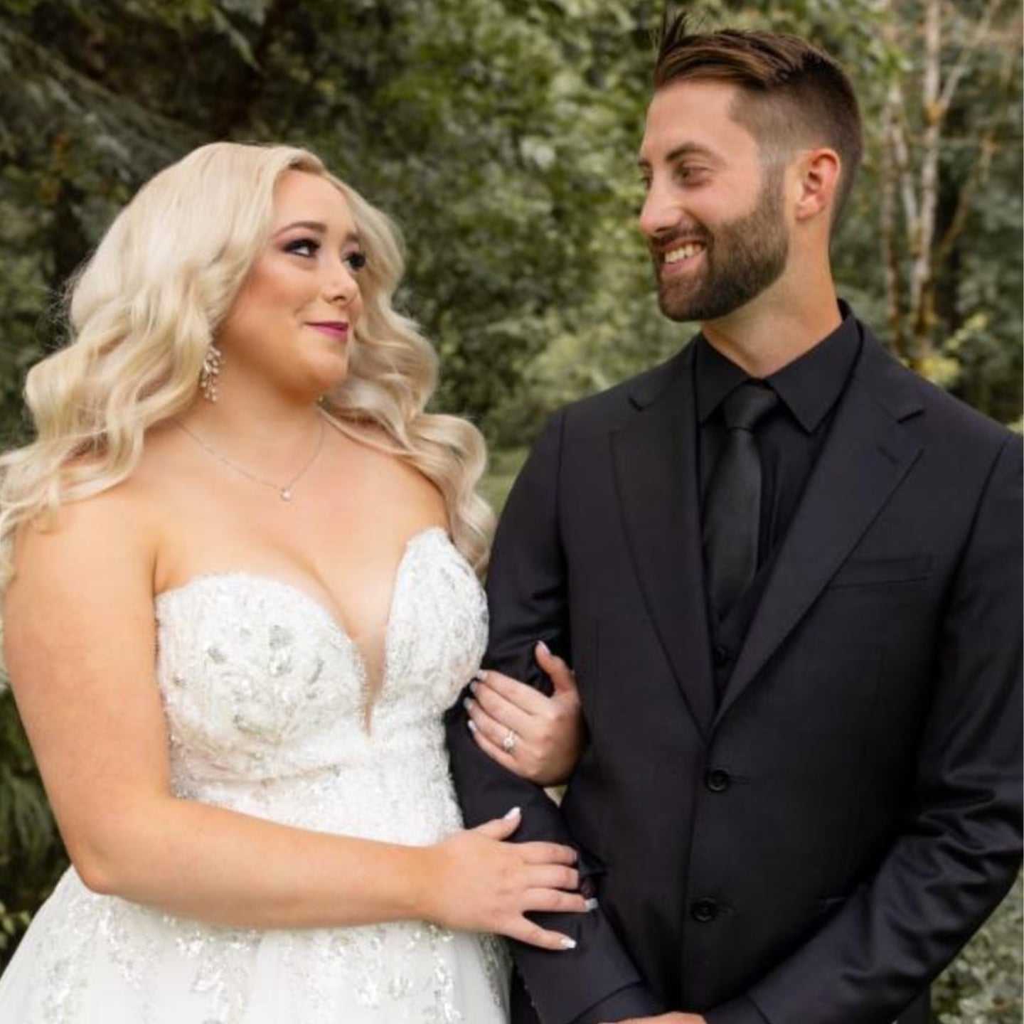 Bride in white dress standing arm-in-arm with groom in black suit, both smiling.