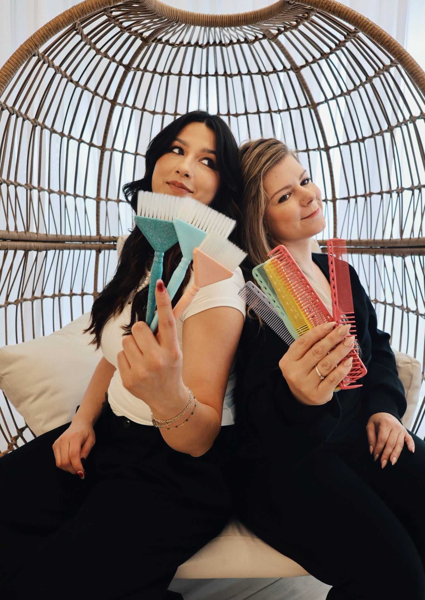 Two women sitting in a wicker chair, holding colorful hairbrushes and smiling.